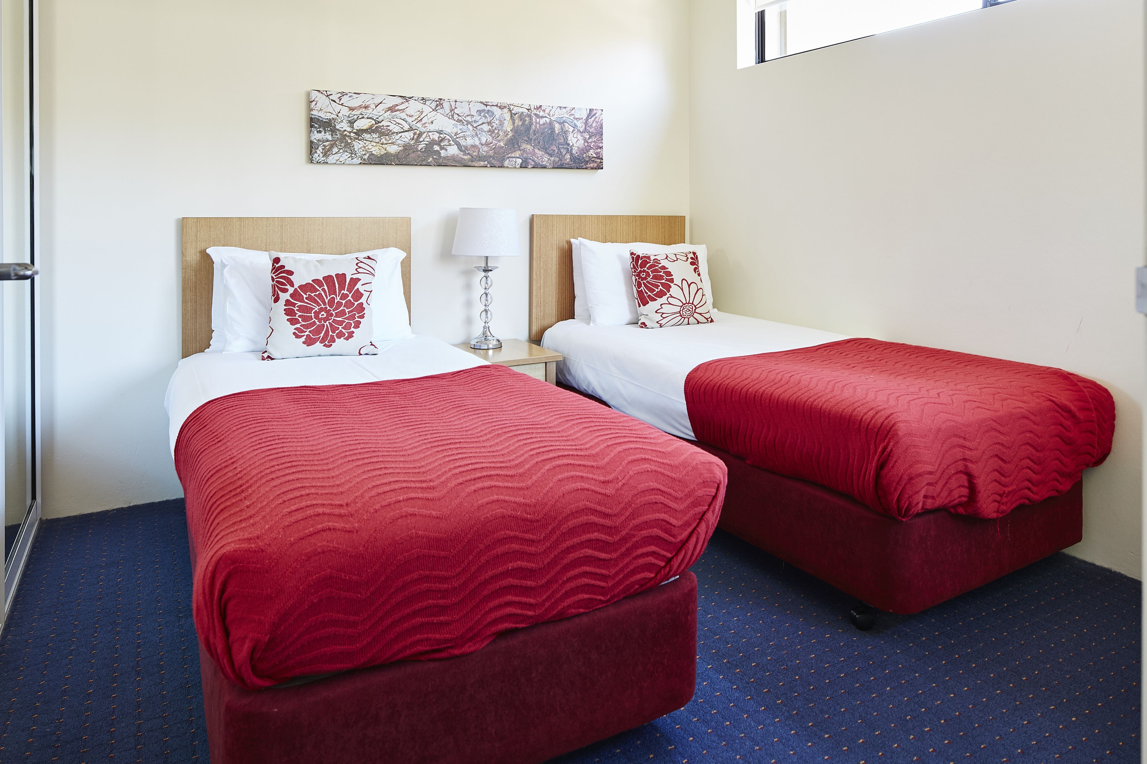 Une chambre d’hôtel avec deux lits, chacun recouvert d’une couverture rouge et agrémenté d’oreillers blancs, une table de chevet avec une lampe et un tableau accroché au mur.