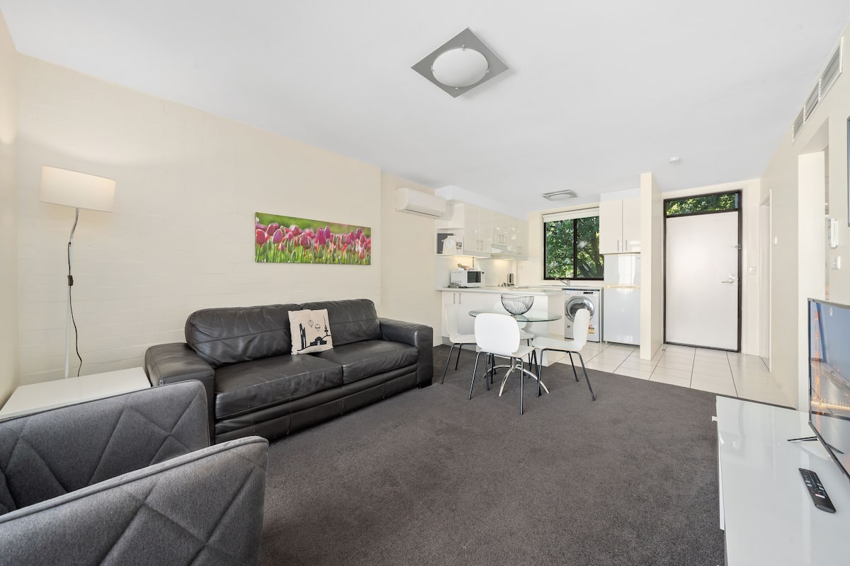 A modern living room with a black leather sofa, a white dining table, and a grey carpet. There is a kitchen area with white cabinets and a washing machine.