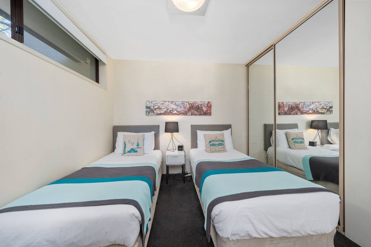 A hotel room with two single beds, a small bedside table with a lamp, a large mirror, and artwork on the wall.