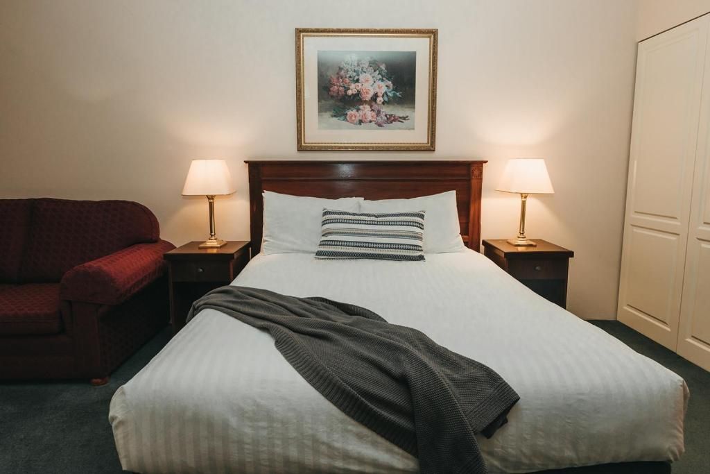 Habitación de hotel con cama, dos lámparas de noche, un cuadro en la pared, un sillón rojo y un armario.