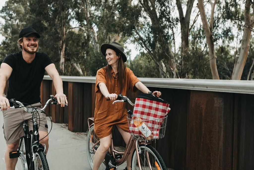 Two people on bicycles with baskets, a bridge, and trees in the background.
