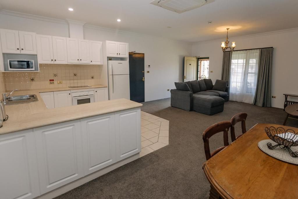 A modern kitchen with white cabinetry, a built-in microwave, and a refrigerator. There is a dining area with a wooden table and chairs, and a sofa in the background.