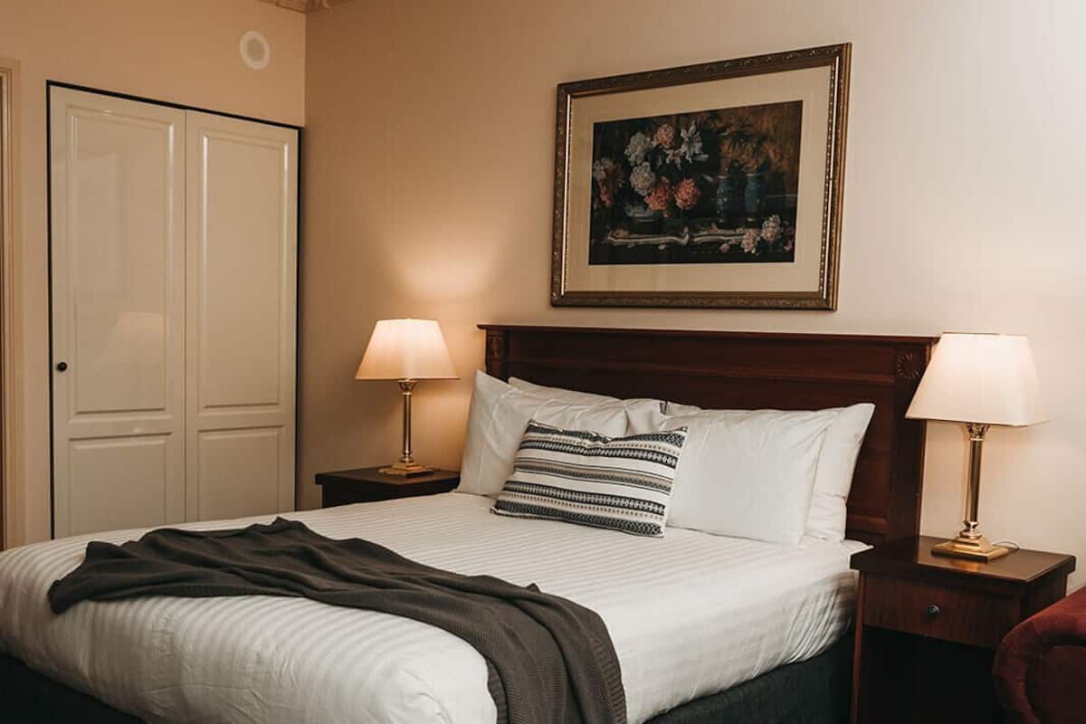 Una cama bien hecha con un cabecero oscuro, dos lámparas de noche, una obra de arte floral enmarcada sobre la cama y una puerta cerrada a la izquierda.