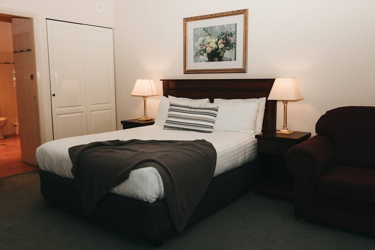 Una cama bien hecha con un cojín estampado, dos lámparas de noche, un cuadro enmarcado sobre la cama y un sillón oscuro a la derecha.