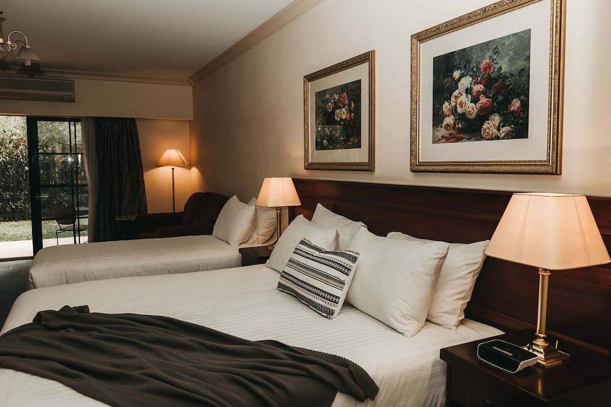 Una habitación de hotel con dos camas, un ventanal grande y dos cuadros enmarcados con motivos florales en la pared.