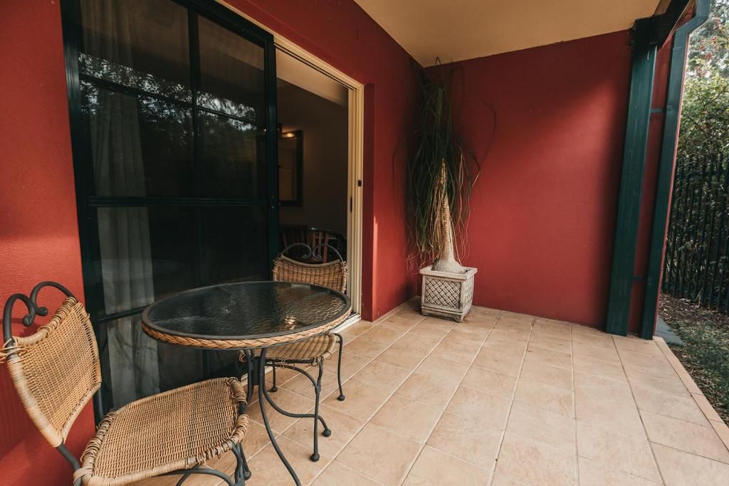 Un patio con una mesa redonda y dos sillas de mimbre, una planta en maceta y un ventanal corredero que da a un espacio interior.