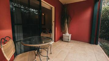 Un patio con una mesa redonda y dos sillas de mimbre, una planta en maceta y un ventanal corredero que da a un espacio interior.