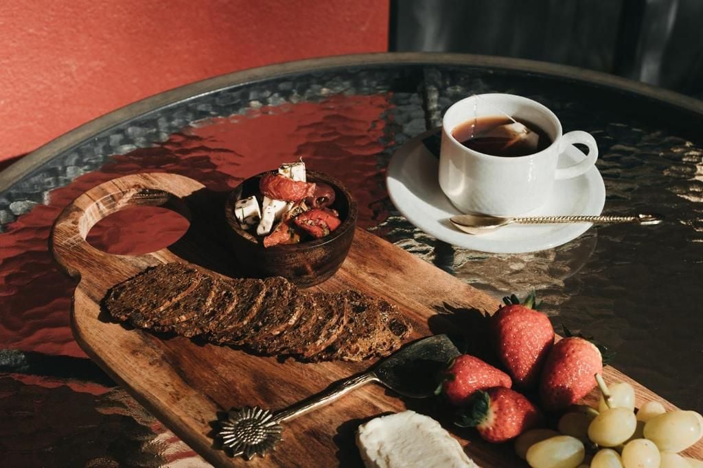 Un tagliere con pane a fette, fragole e uva, una ciotola di dessert e una tazza di caffè su un tavolino di vetro.