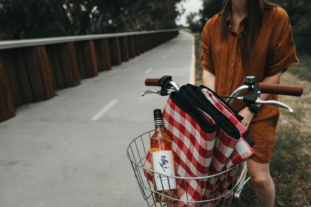 A person riding a bicycle with a basket containing a bottle and a checkered cloth.