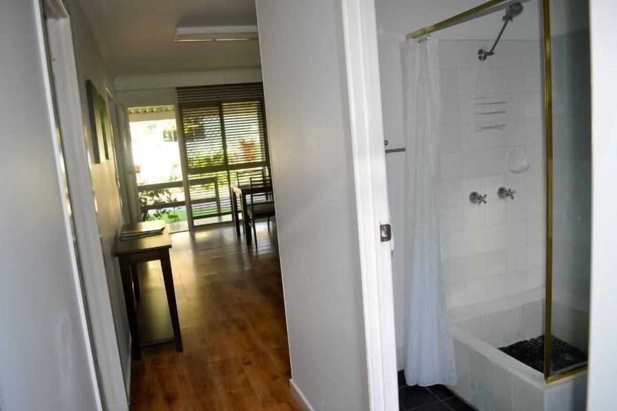 A hallway leading to a bathroom with a glass shower enclosure and a view of a balcony with a table and chairs.