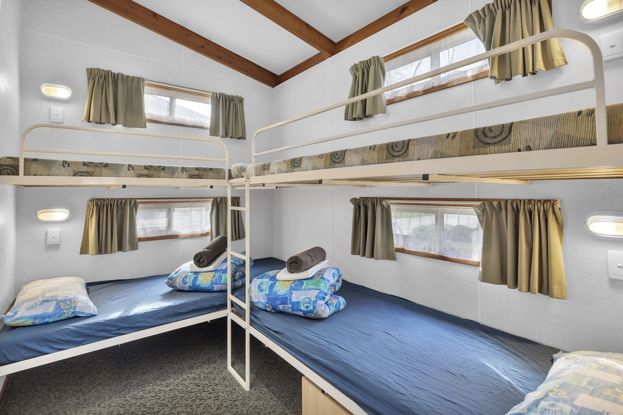 A bunk bed room with two beds, each with a pillow and a blanket. There are windows with curtains, and a ceiling light is visible.