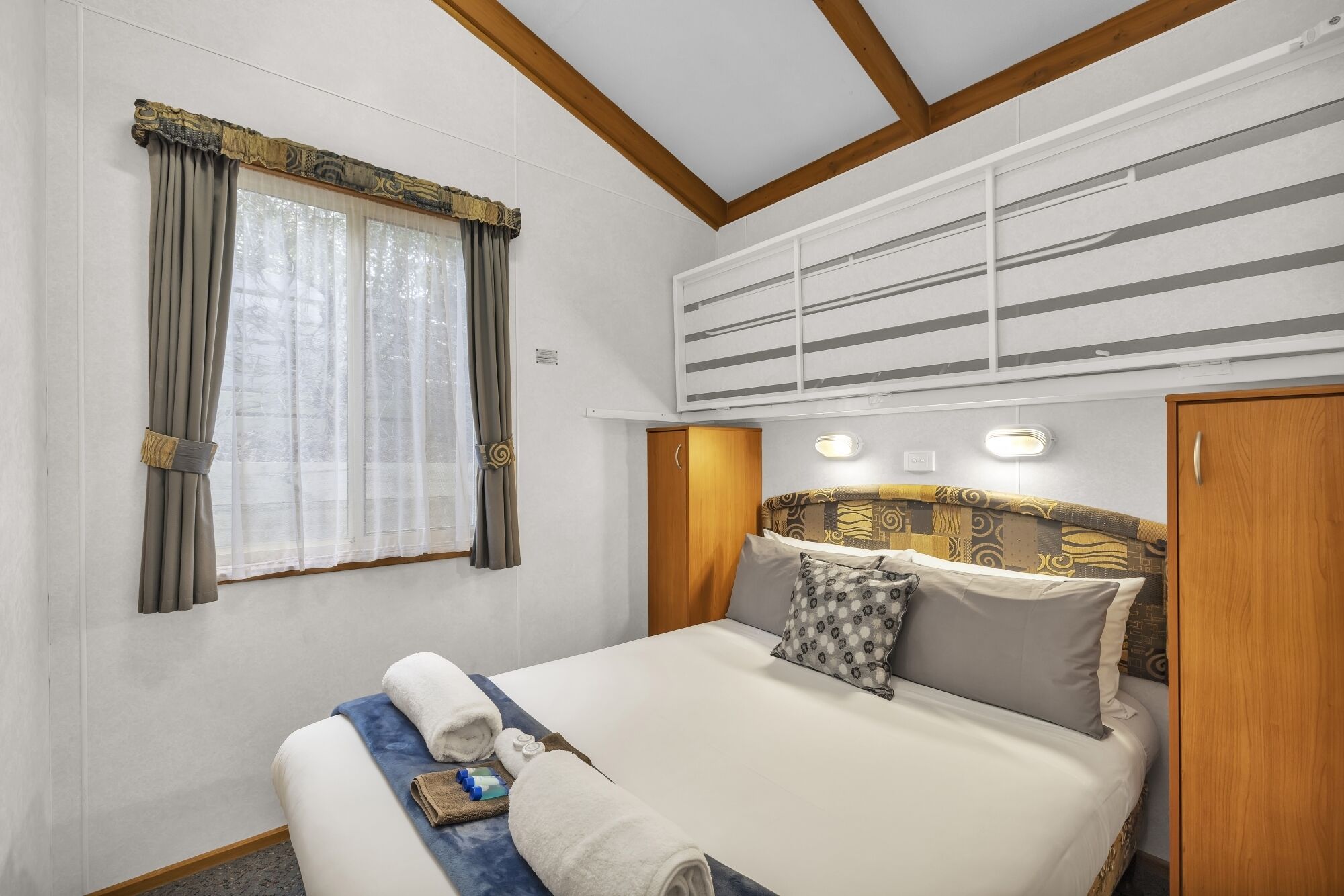 Una cama bien hecha con cabecera, una ventana con cortinas y una lámpara fijada a la pared.