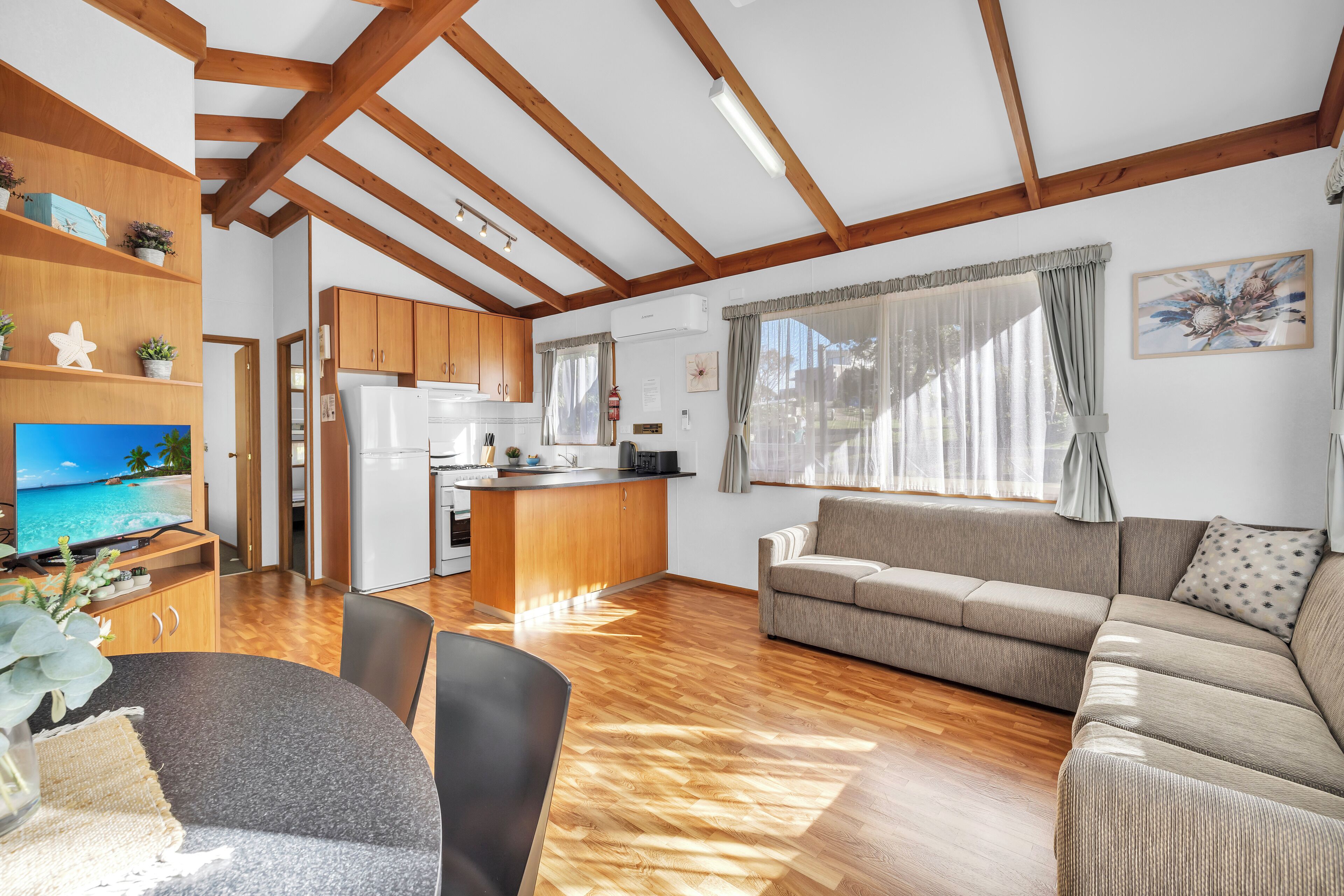 A spacious living area with wooden beams, a kitchen, a sofa, and a TV.