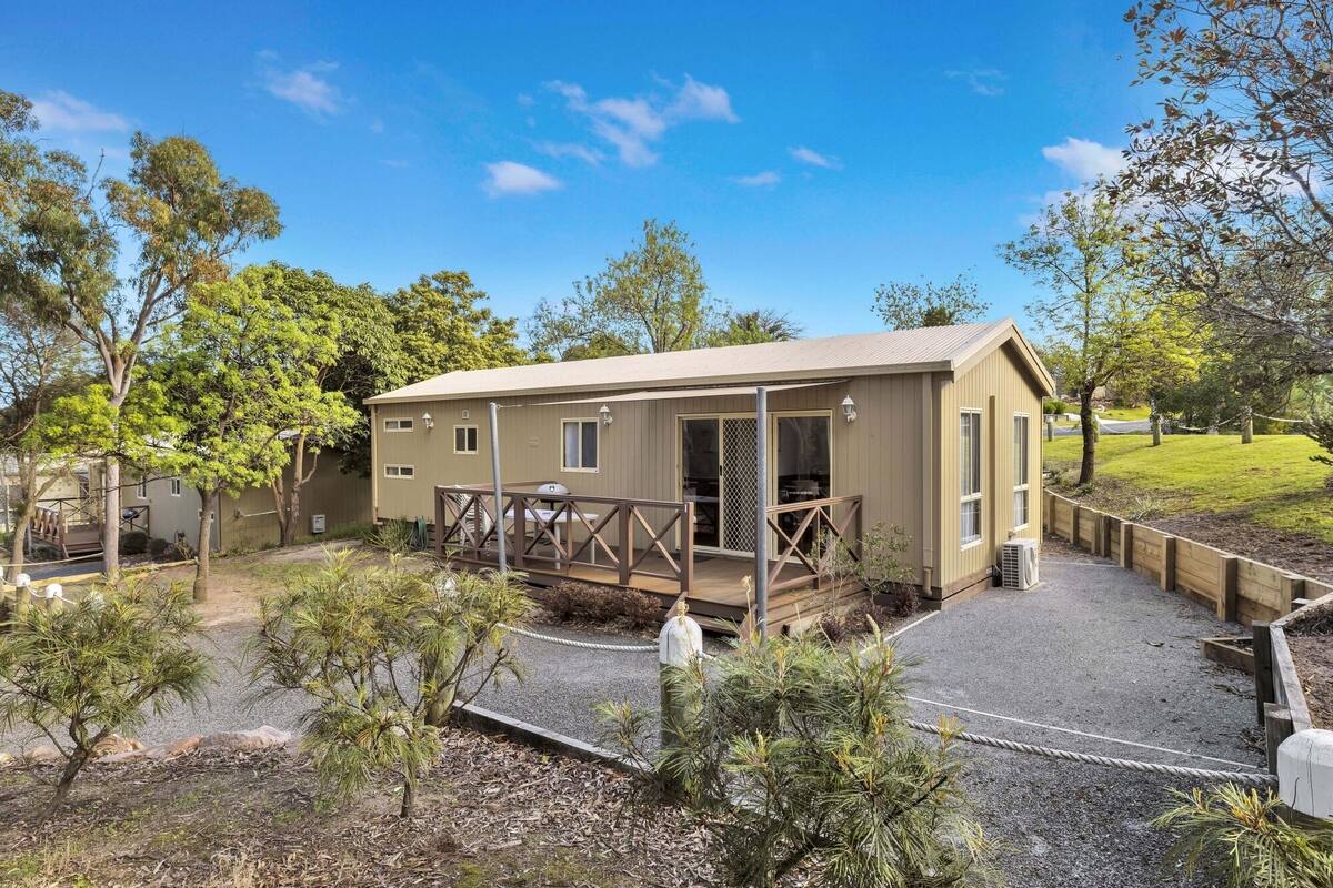 Une maison mobile d’un seul étage, de couleur beige, dotée d’un patio couvert et entourée d’un jardin aménagé avec différents arbustes et arbres.