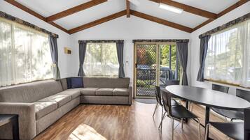 Una habitación con sofá modular, una mesa de comedor con sillas, piso de madera y ventanas grandes con cortinas.
