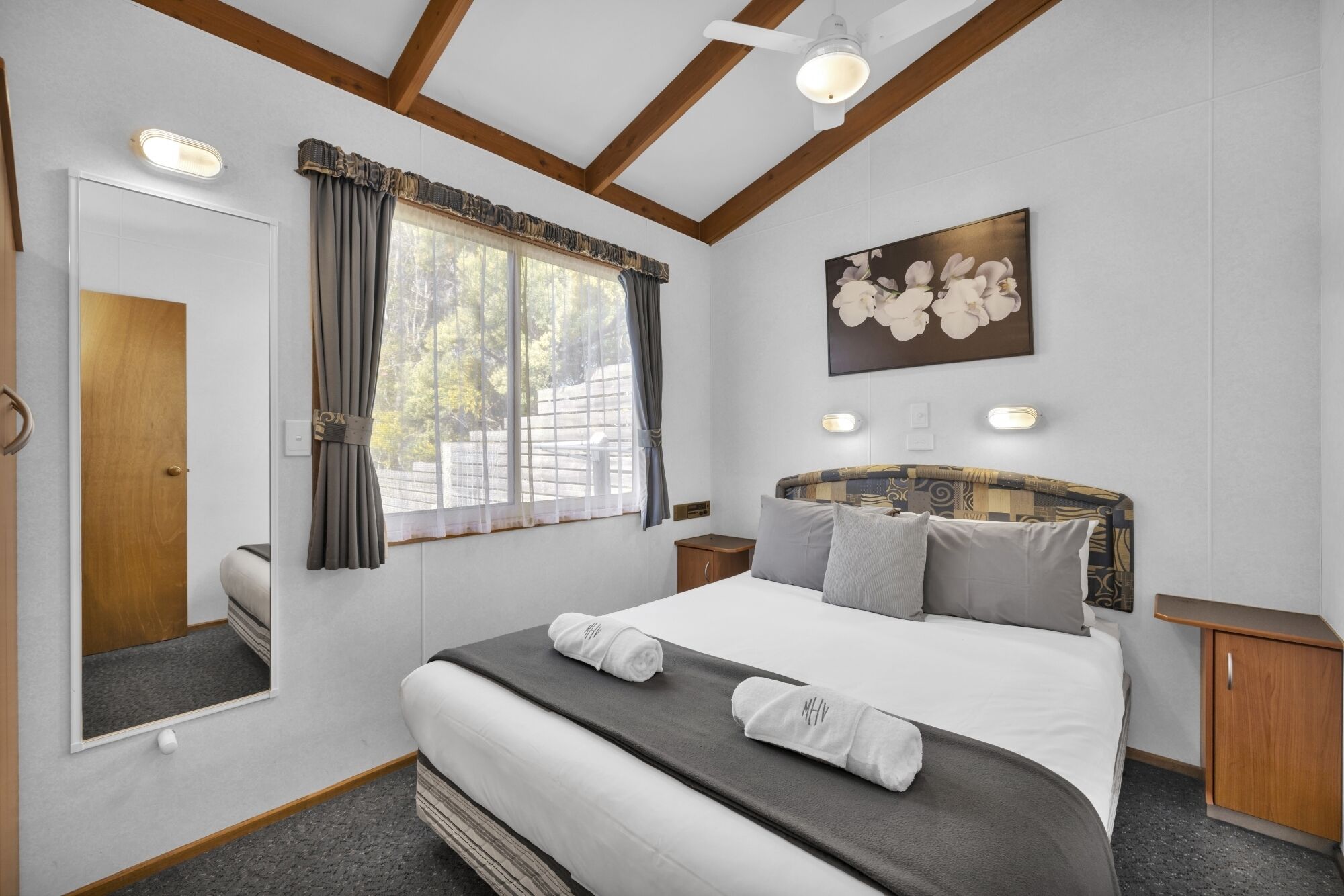 Une chambre à coucher avec un grand lit, une tête de lit ornée d’un motif floral, une fenêtre avec des rideaux, un miroir et un tableau accroché au mur.