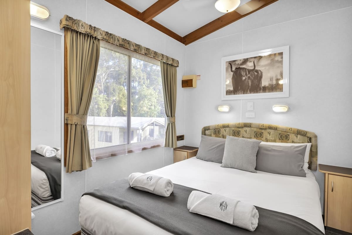 Une chambre d’hôtel avec un grand lit, une fenêtre avec des rideaux, une tête de lit ornée d’un motif, un tableau encadré au mur et une table de chevet avec une lampe.
