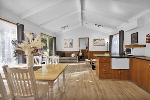 Amplia habitación con una mesa de comedor de madera, sillas blancas, un sofá y una cocina con armarios de madera y un microondas.