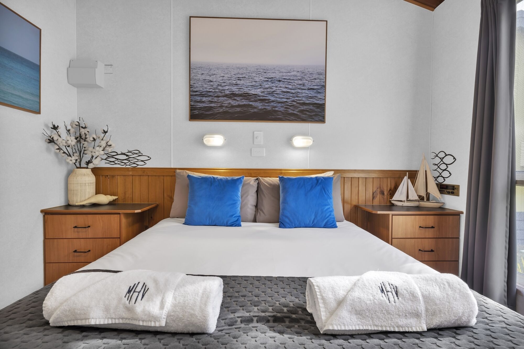 Une chambre d’hôtel avec un lit, deux tables de chevet, une tête de lit en bois, un tableau représentant la mer accroché au mur, un vase avec des branches et une maquette de bateau.