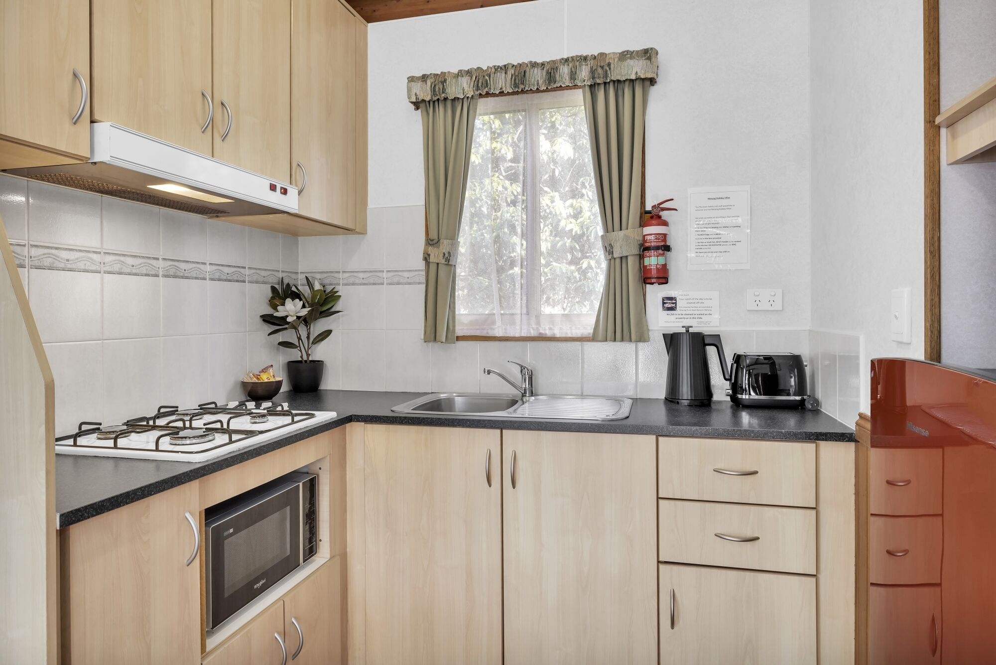 Une cuisine de taille réduite équipée d’une cuisinière à gaz, d’un four à micro-ondes, d’un évier et d’une fenêtre avec des rideaux.