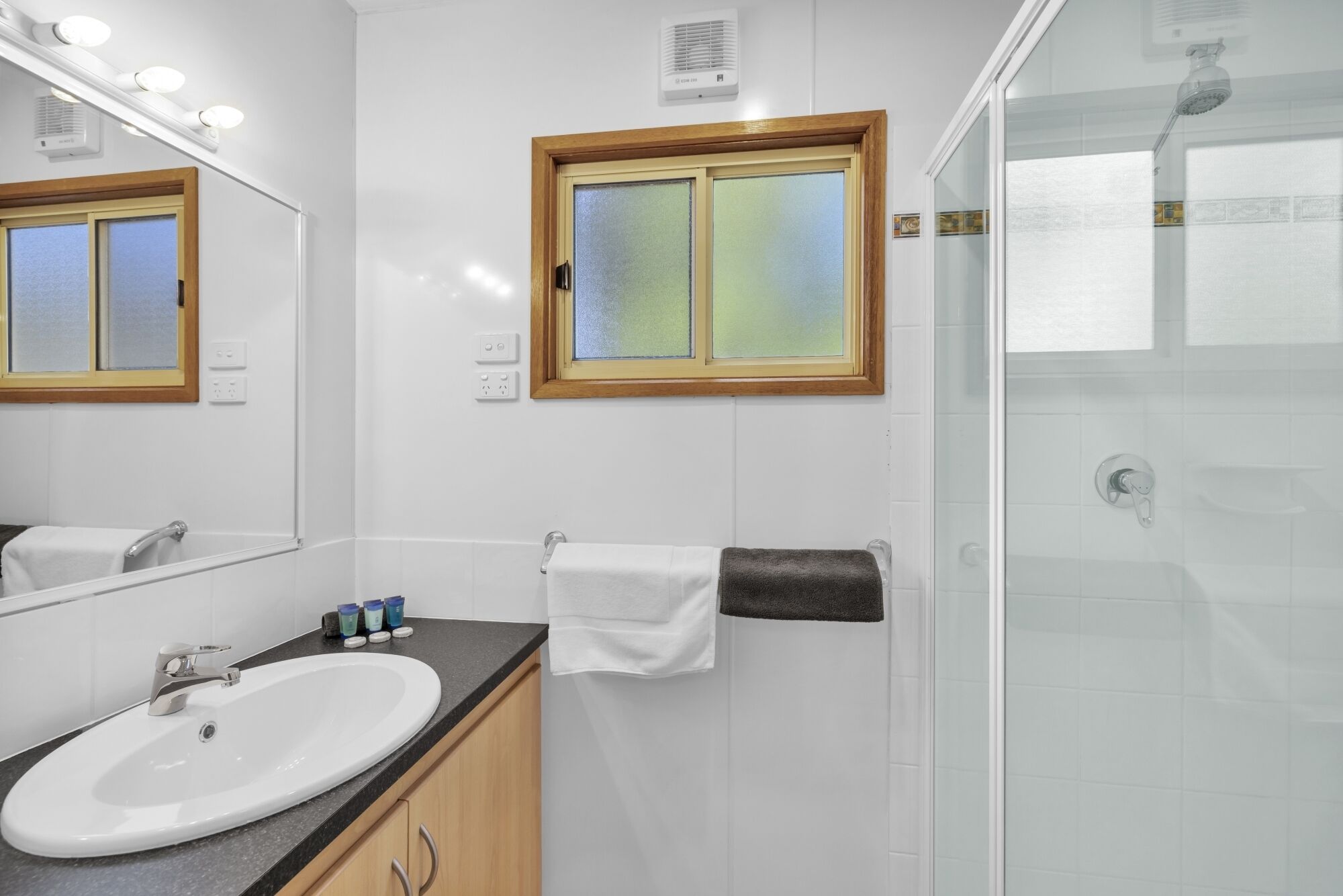 Une salle de bain avec un lavabo blanc, un miroir, une fenêtre à verre dépoli et un espace douche.