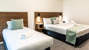 Een hotelkamer met twee eenpersoonsbedden, elk met een houten hoofdbord, wit beddengoed en een groene plaid. Er staan nachtkastjes met lampen en een klein plankje.