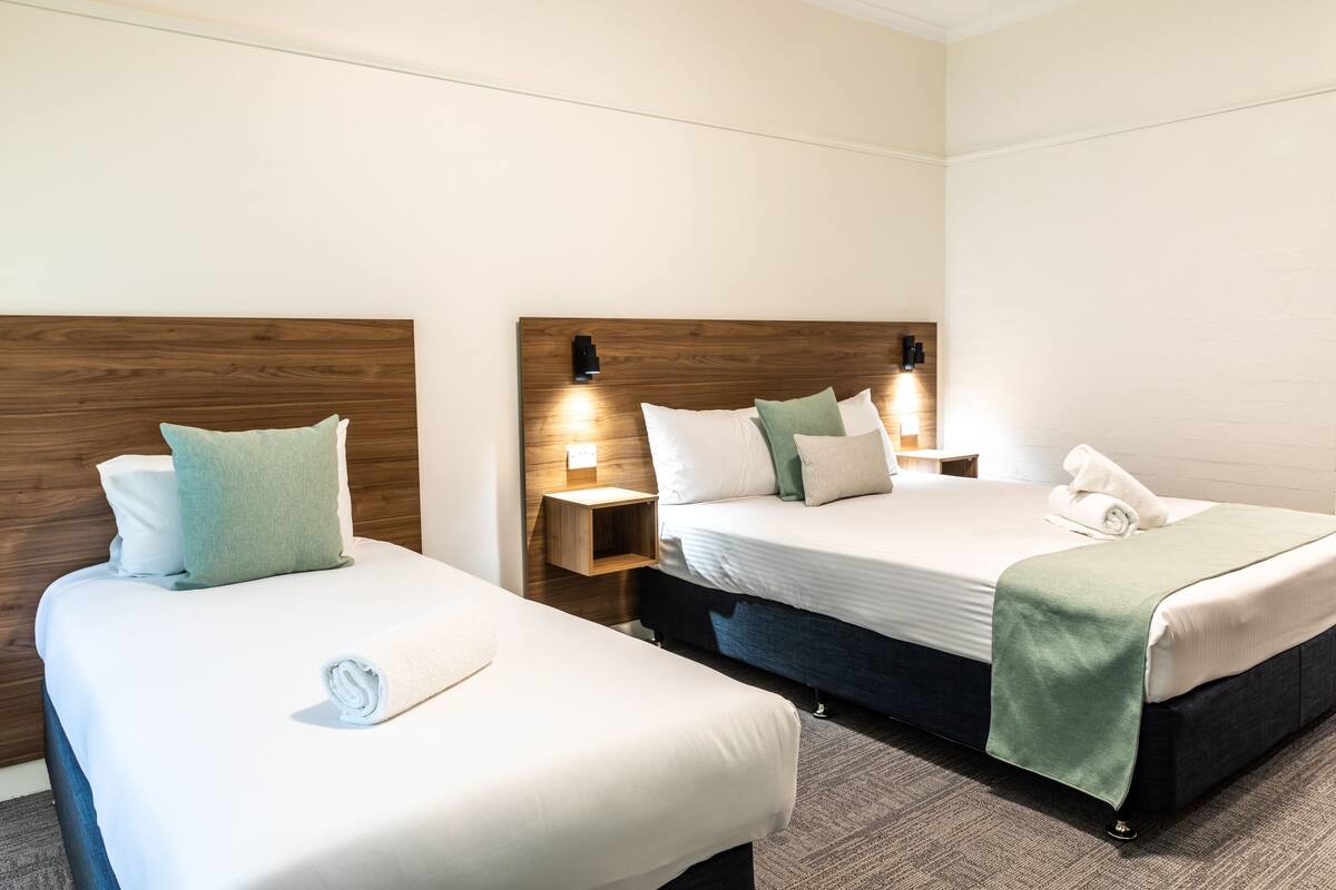 A hotel room with two single beds, each with a wooden headboard, white bedding, and a green throw blanket. There are bedside tables with lamps and a small shelf.