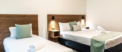 Una habitación de hotel con dos camas individuales, cada una con cabecero de madera, ropa de cama blanca y una manta verde. Hay mesitas de noche con lámparas y un estante pequeño.