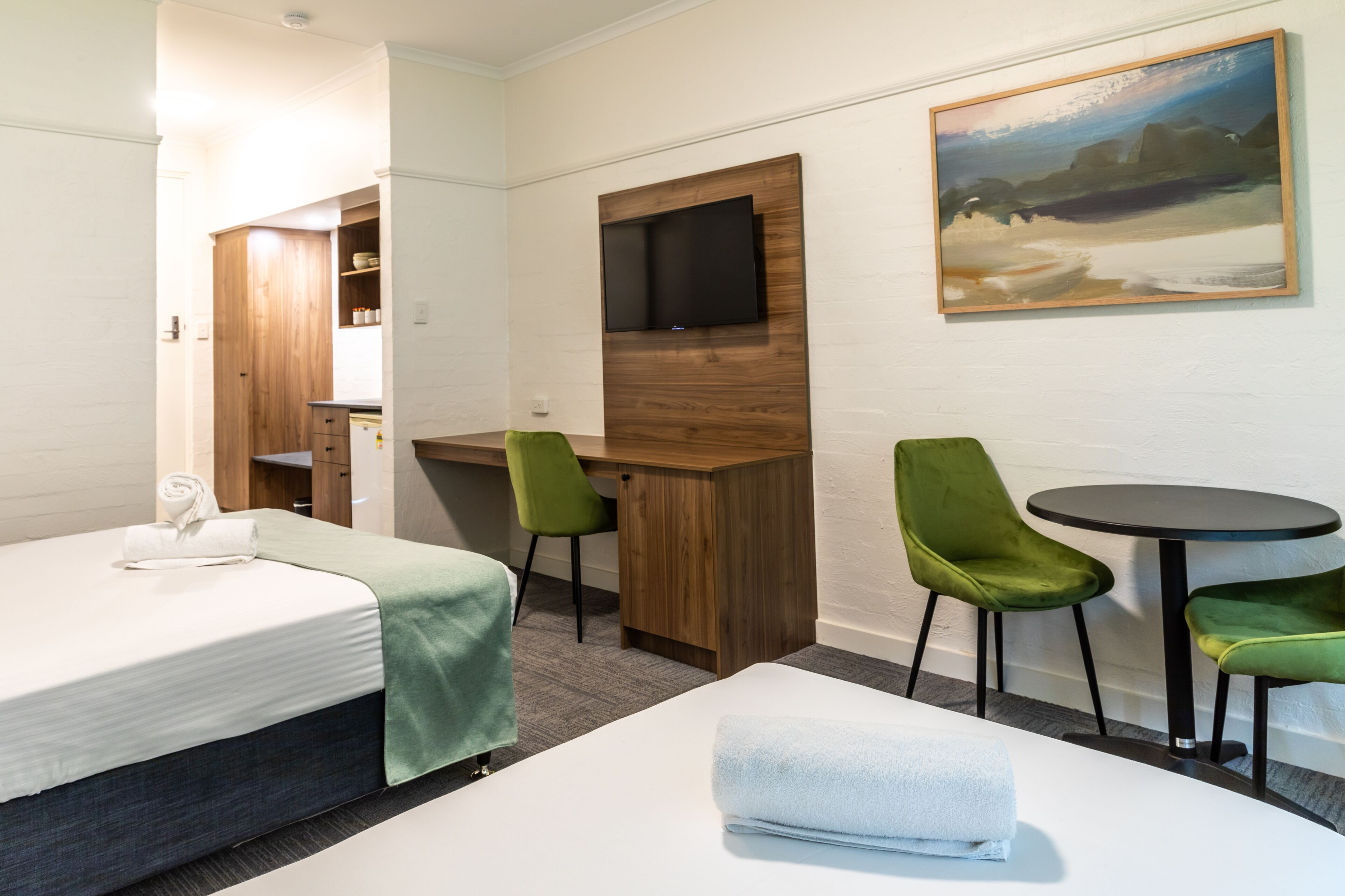 A hotel room with a bed, a desk, two green chairs, a small table, a TV mounted on a wooden panel, and a painting on the wall.