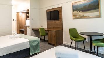 A hotel room with a bed, a desk, two green chairs, a small table, a TV mounted on a wooden panel, and a painting on the wall.