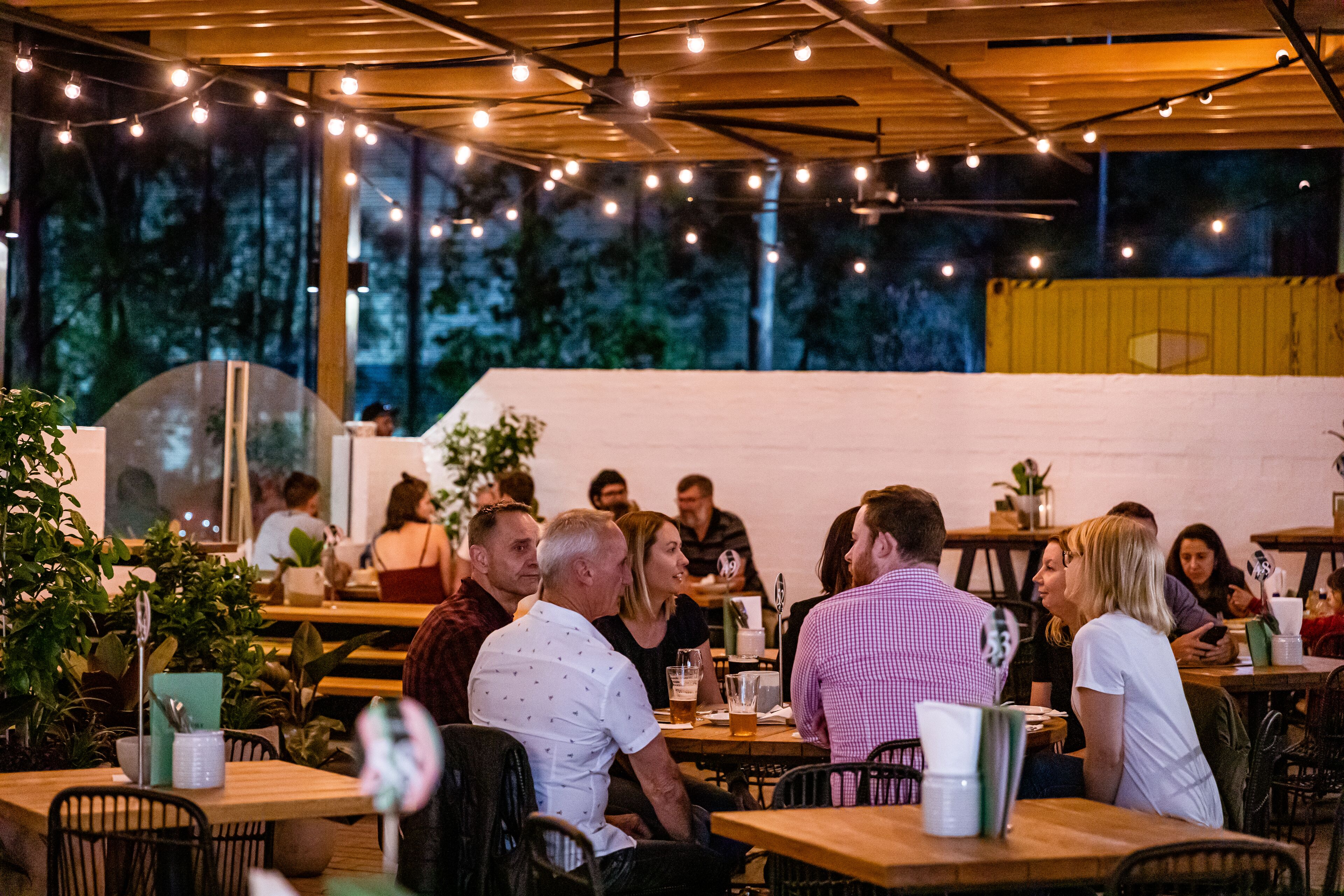 An outdoor dining area with wooden tables and chairs, string lights overhead, and a view of trees through large windows.