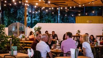 An outdoor dining area with wooden tables and chairs, string lights overhead, and a view of trees through large windows.