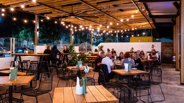 Een terras met houten tafels en stoelen, sfeerverlichting erboven en een uitzicht op de straat op de achtergrond.