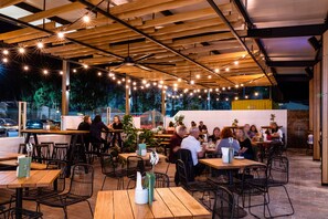 Un espace de restauration en plein air avec des tables et des chaises en bois, des guirlandes lumineuses suspendues et une vue sur la rue en arrière-plan.