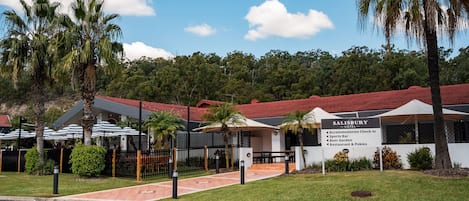 Un hotel con techo rojo, palmeras y un cartel que enumera servicios como alojamiento, bar deportivo, cervecería, restaurante y máquinas tragamonedas.