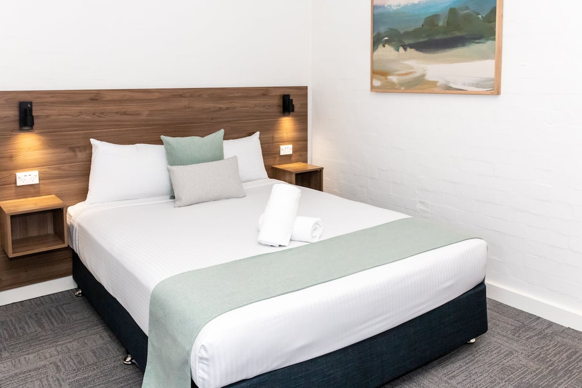 A neatly made bed with a green throw blanket, white bedding, and multiple pillows. There are two bedside tables with lamps, a wall-mounted light, and a painting above the bed.