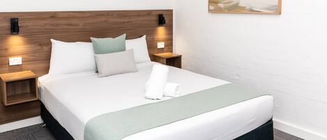 Una cama bien hecha con una manta verde, ropa de cama blanca y varias almohadas. Hay dos mesitas de noche con lámparas, una luz en la pared y un cuadro sobre la cama.