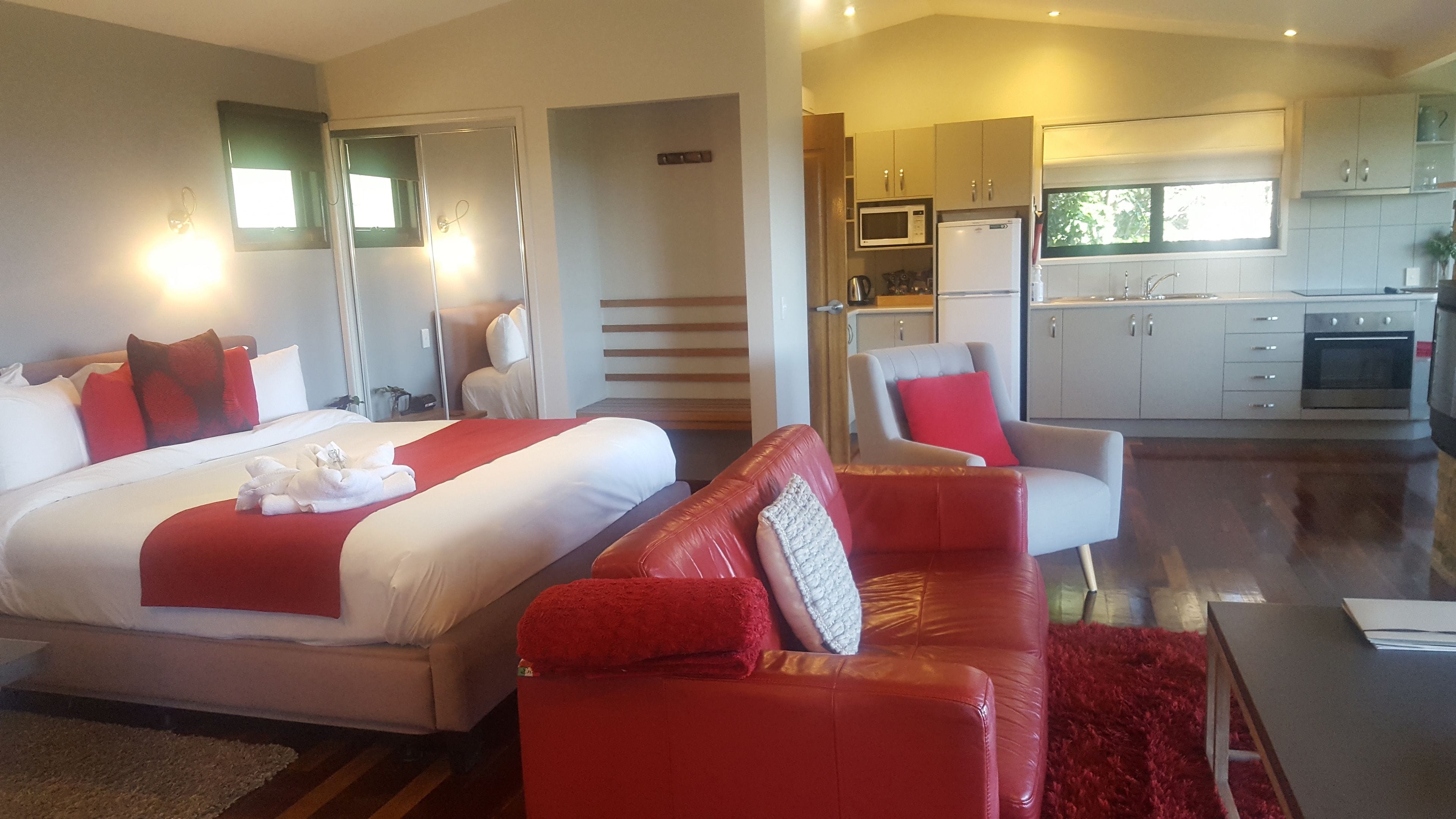 Habitación de hotel con una cama grande, un sofá rojo, una zona de cocina con microondas y horno, y vistas al exterior.