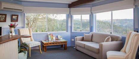 Ein gemütliches Zimmer mit einem Holztisch, einem beige-farbenen Sofa und einem gestreiften Sessel. Ein Holztisch bietet Platz für Bücher und einen Blumentopf. Große Fenster bieten Blick ins Freie.
