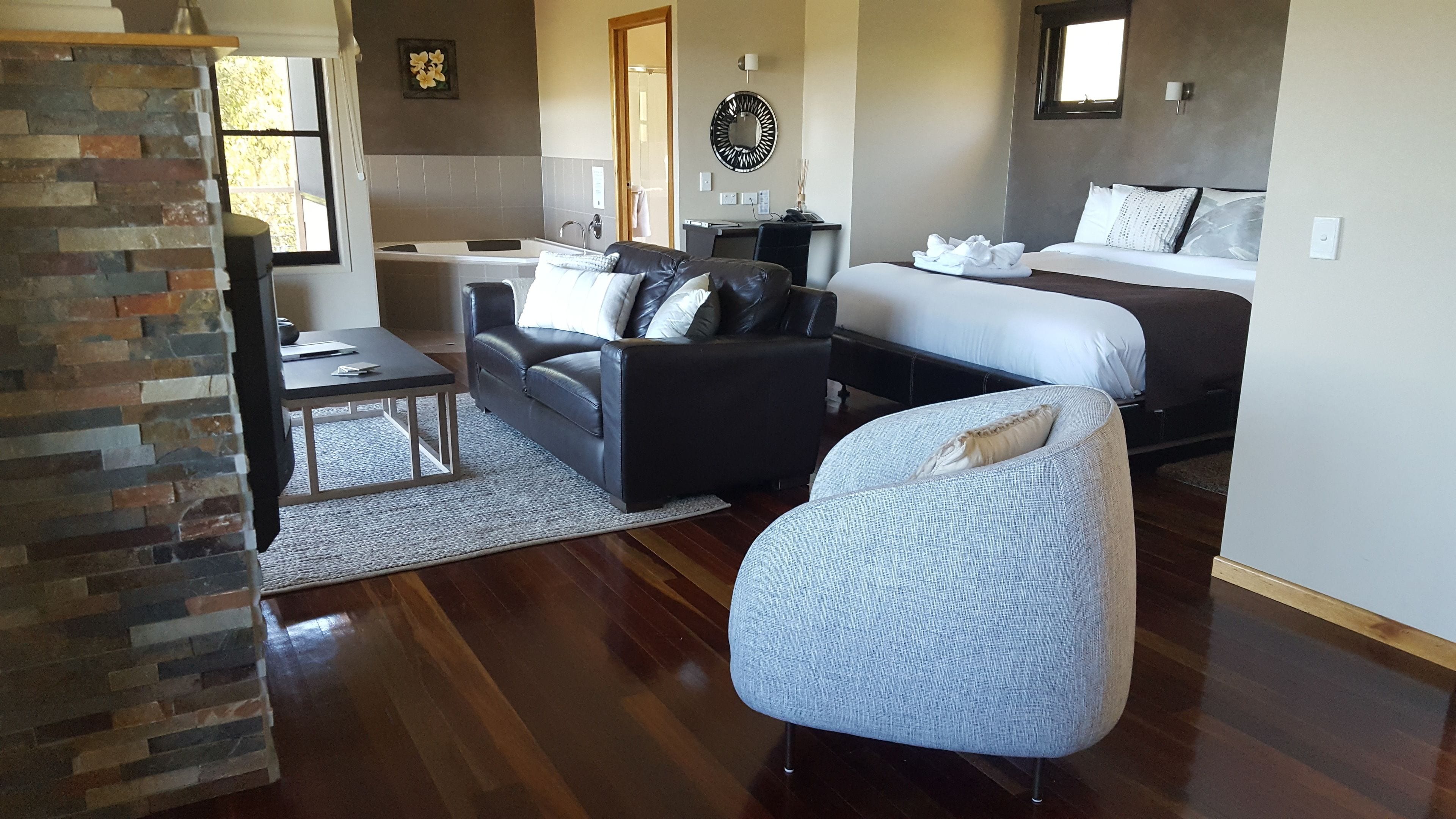 A modern living room with a stone accent wall, a black leather sofa, a grey armchair, a wooden coffee table, and a large window.