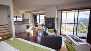 Ampla sala de estar com uma cama grande, um sofá e uma lareira. Cozinha visível ao fundo, com vista para as montanhas.