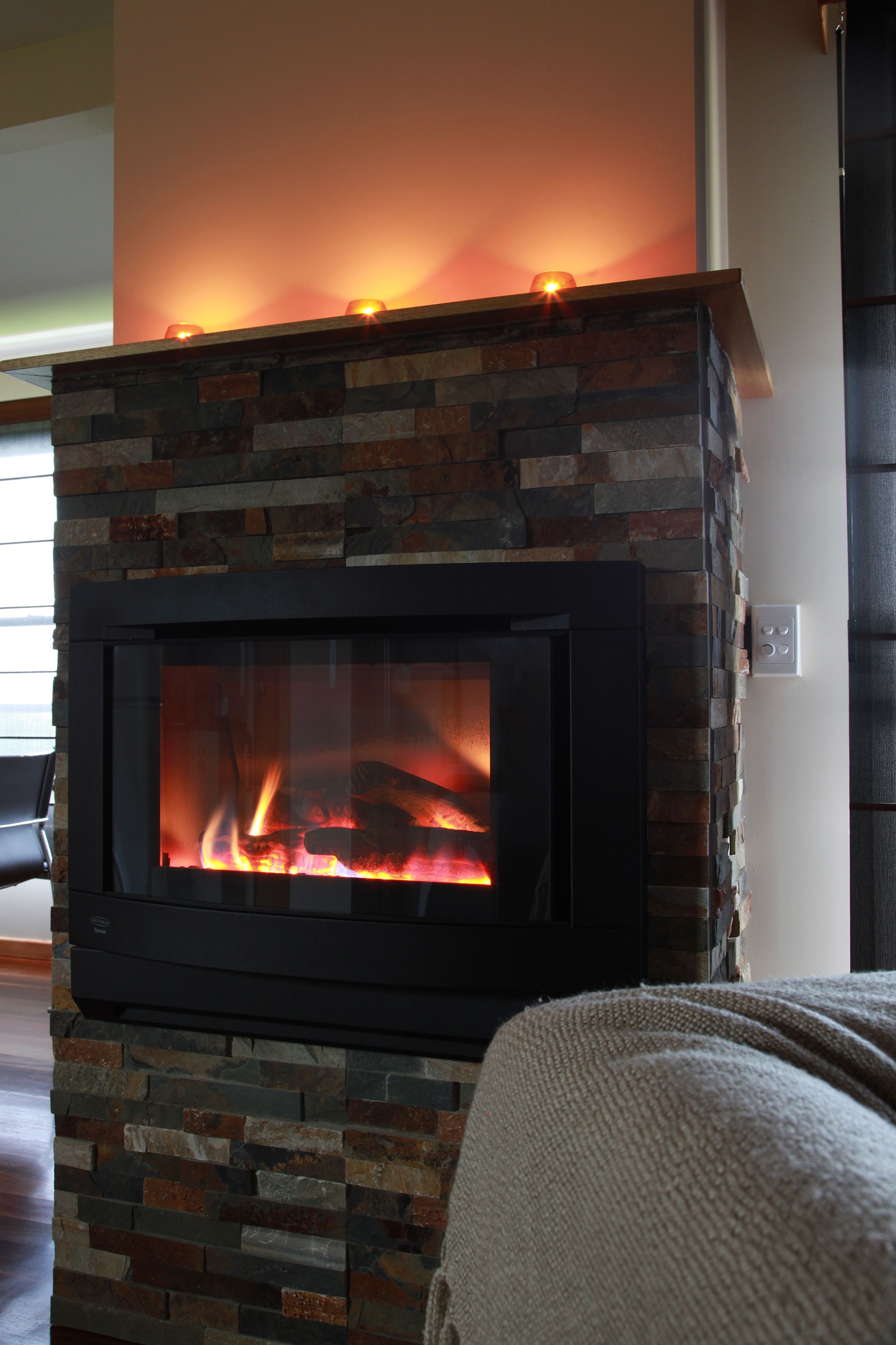 A modern fireplace with a stone facade and a glass door, set in a living room with a sofa and a wooden floor.