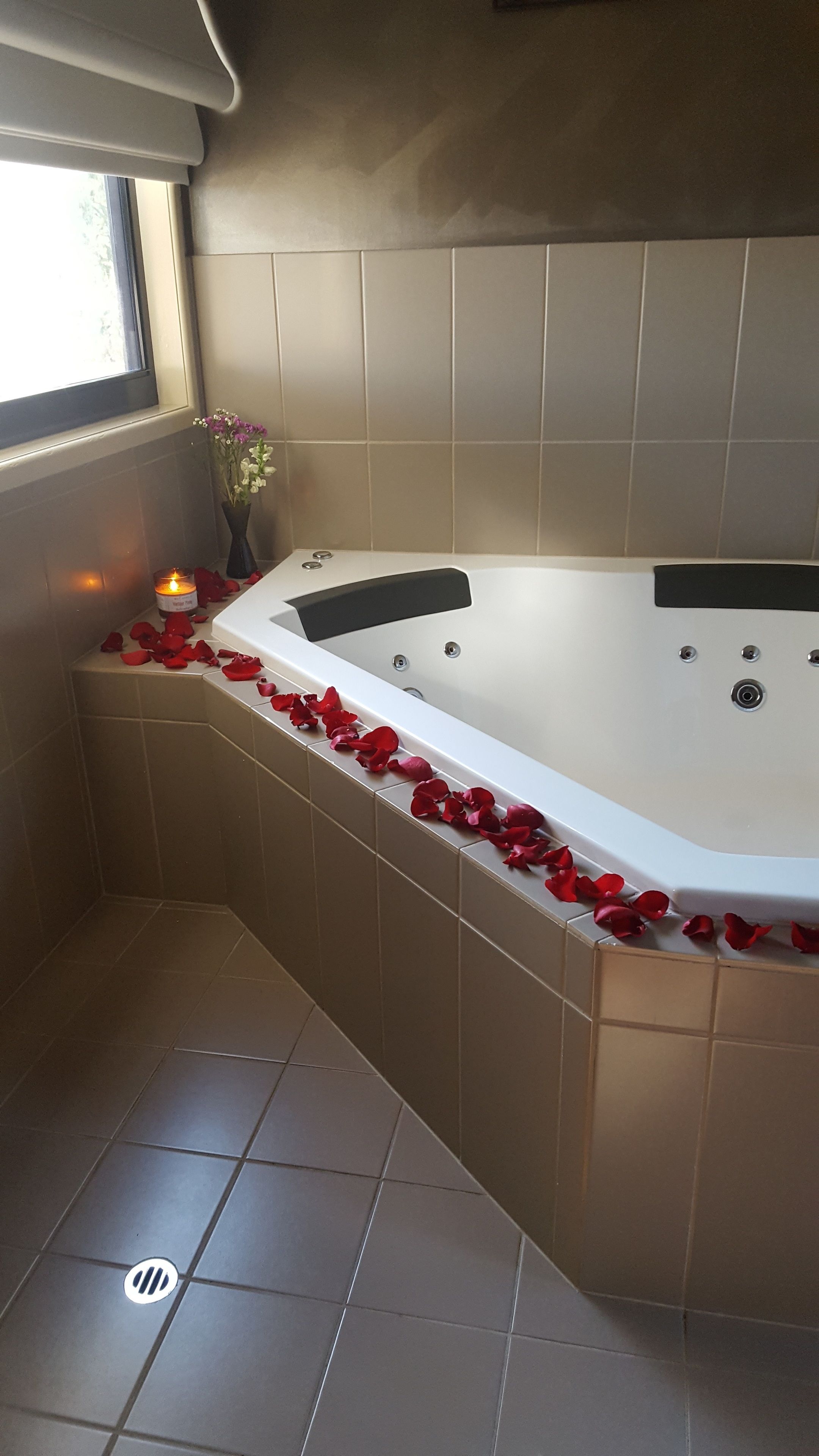 A corner bathroom with a bathtub, tiled walls, a window, and a vase with flowers.