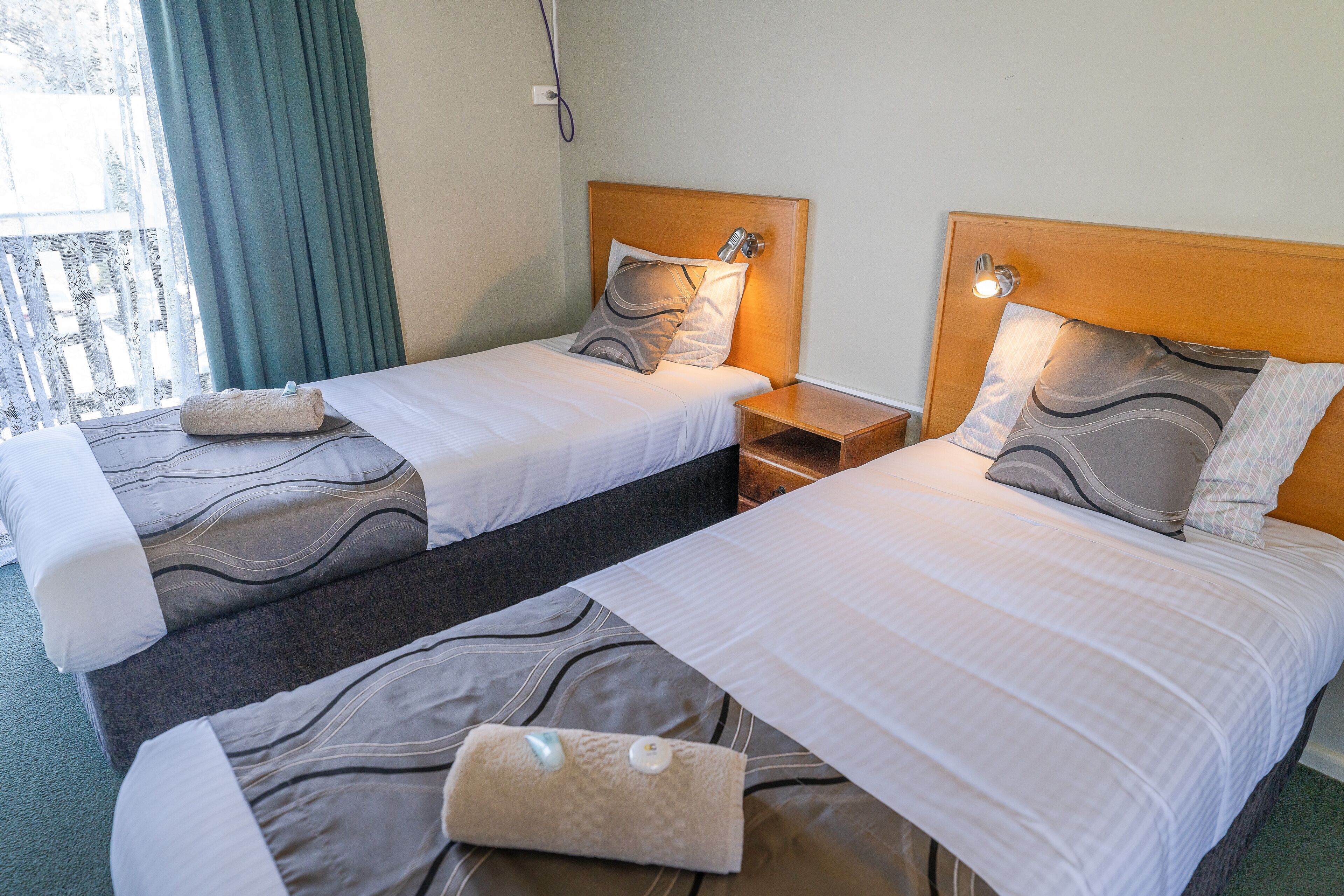 Two single beds with wooden headboards, a small bedside table, and a window with curtains.