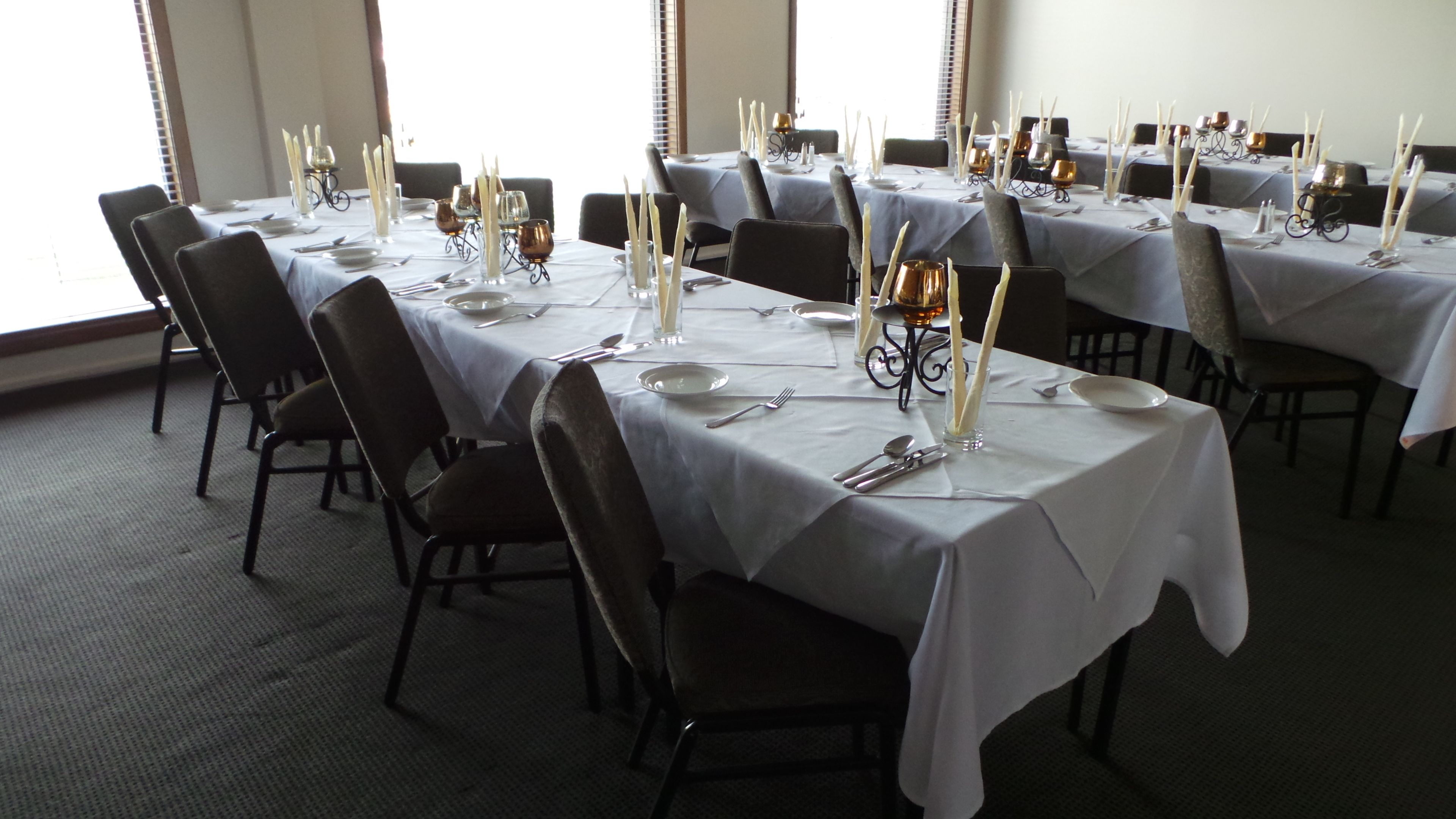 A dining area with tables set for a meal, featuring white tablecloths, dark chairs, and candle holders.