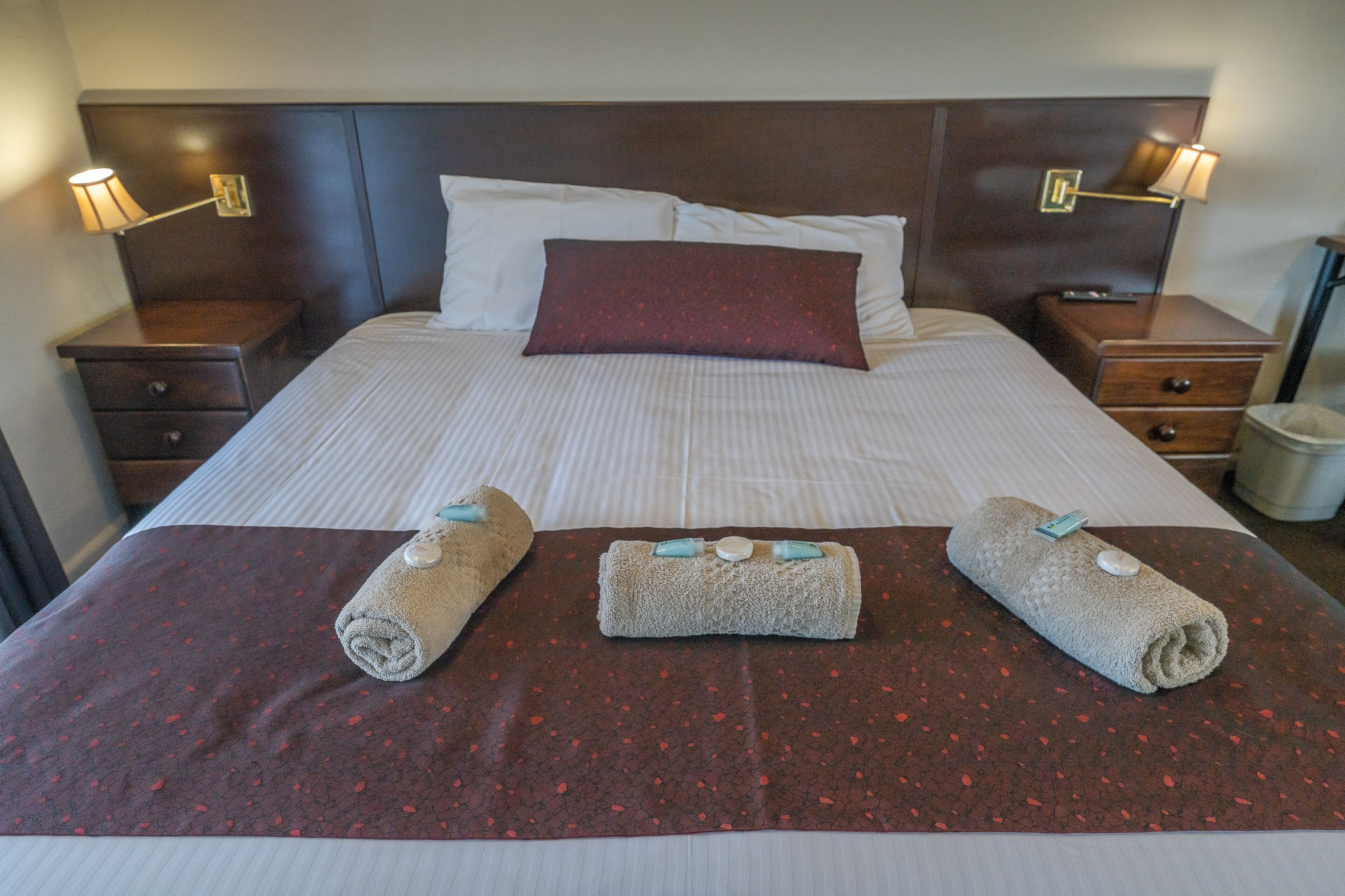 A neatly made bed with a red patterned bedspread, two rolled towels with blue bows, and two bedside lamps.