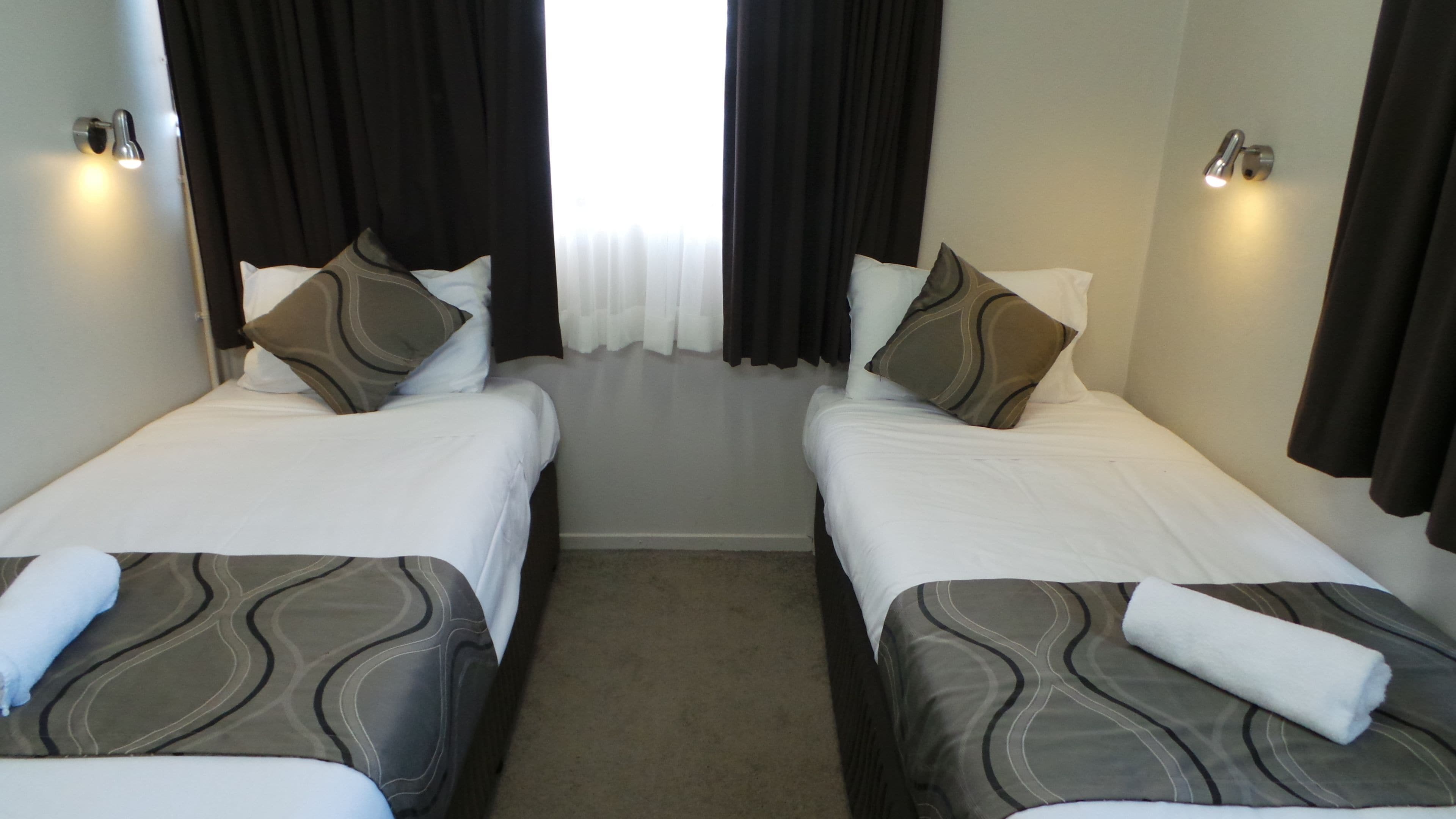 A hotel room with two single beds, each with a patterned bedspread and pillows, a carpeted floor, and a window with curtains.