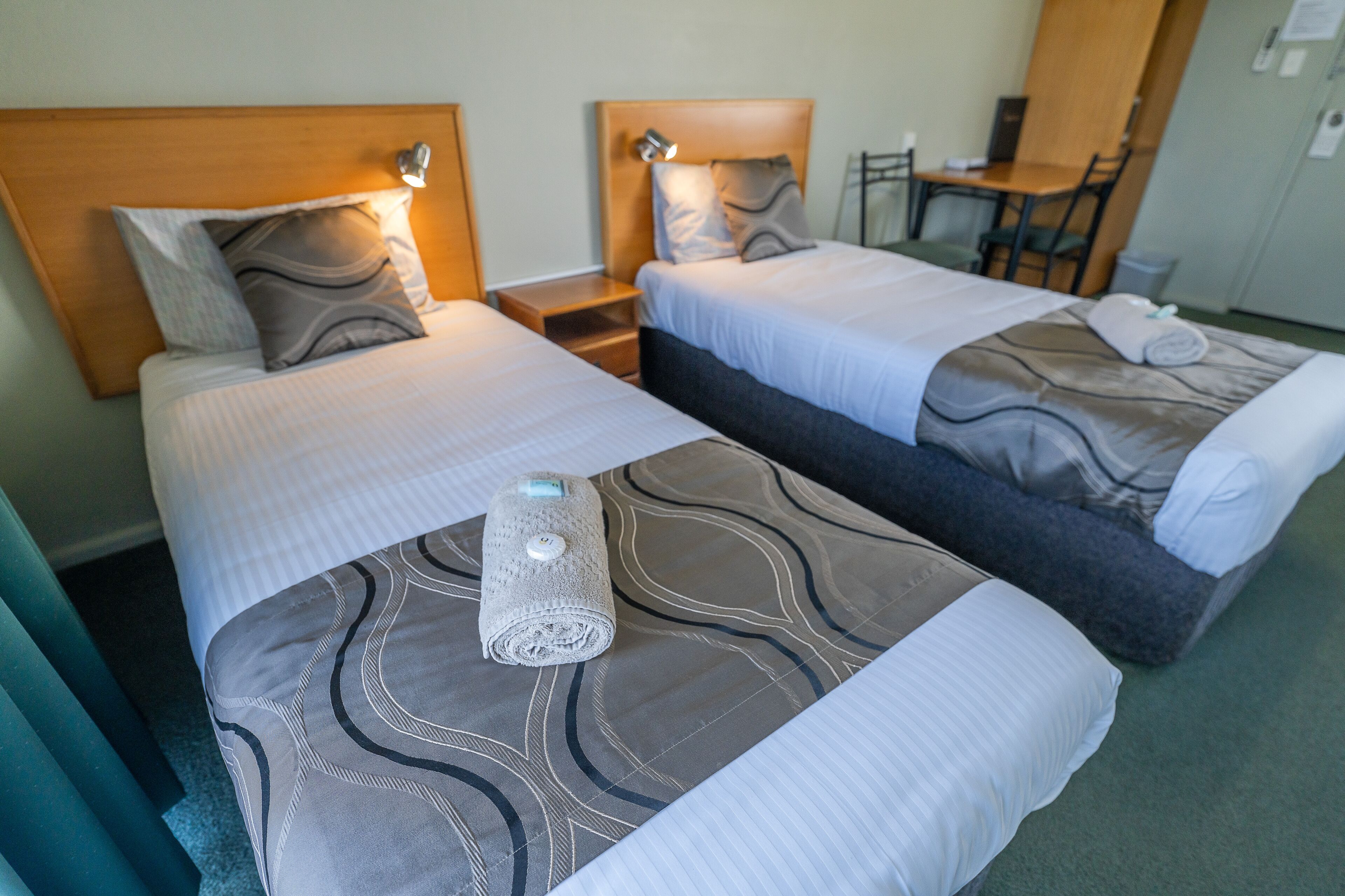 Quarto de hotel com duas camas, cada uma com cabeceira e mesas de cabeceira. Há uma pequena mesa e uma cadeira no canto.