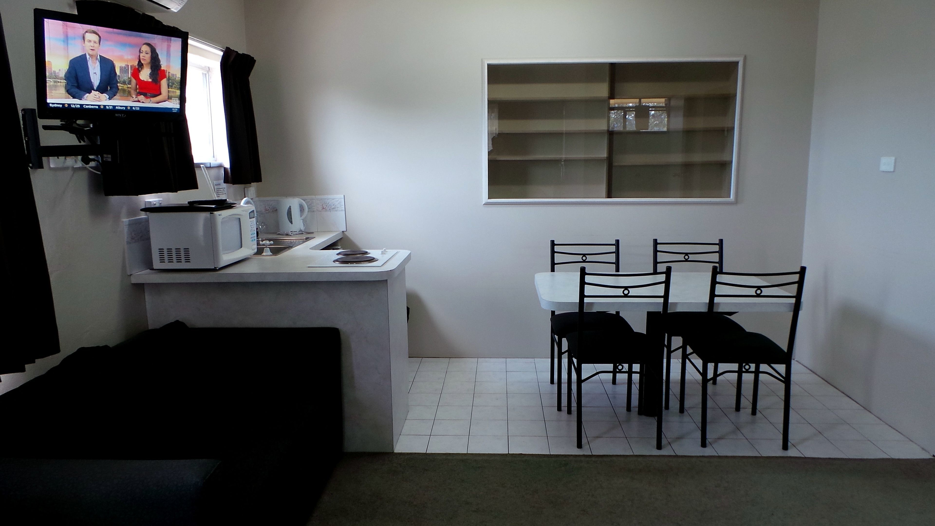 A small, modern kitchen with a microwave, kettle, and coffee maker. There is a dining area with a table and four chairs. A television is mounted on the wall displaying a news program.