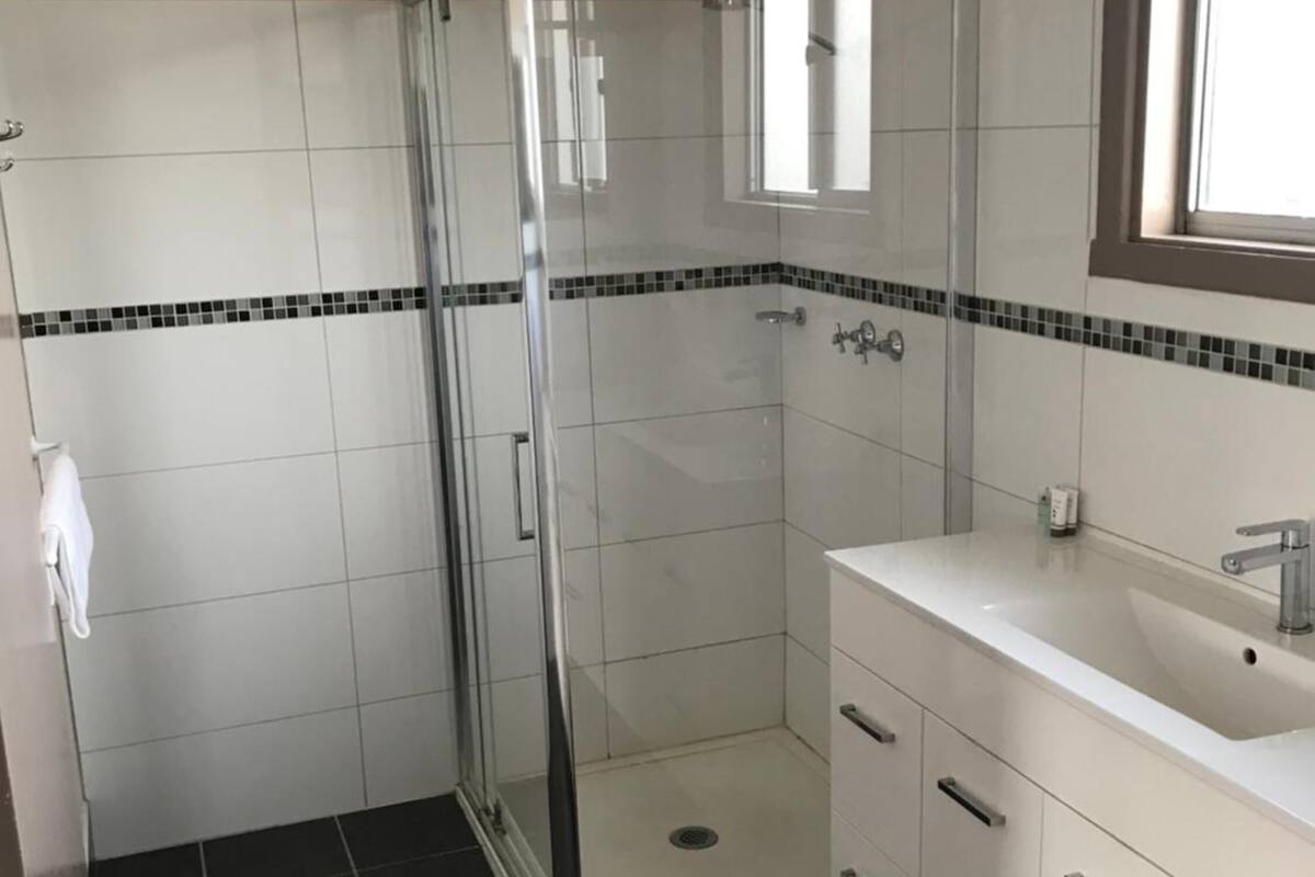 Ein Badezimmer mit einer gläsernen Duschkabine, weiß gefliesten Wänden, einer schwarz-weißen Fliesergrenze, einem weißen Waschbecken mit Spiegel darüber und dunklen Bodenfliesen.